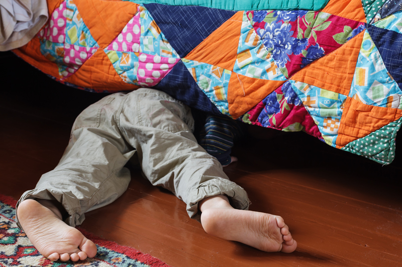 a small boy searches for something under his bed. All that is visible is just his legs and feet from under the bed with a very colourful multi patterned quilt.